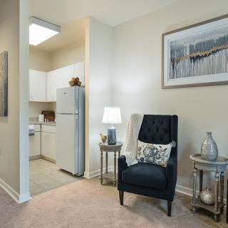 A cozy living space adjacent to a small kitchen area, featuring a comfortable black chair, side tables with decor items, and wall artwork.