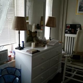 A cozy bedroom with a white dresser and two lamps in front of a window with blinds.