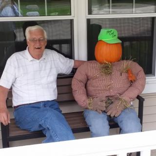 A man sitting on a porch bench next to a scarecrow with a pumpkin head.
