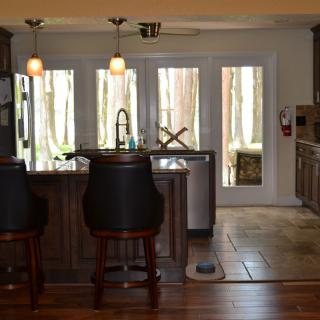 A modern kitchen interior featuring bar stools, wooden cabinets, and large windows offering natural light.