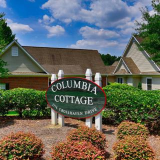 Entrance sign of Columbia Cottage surrounded by greenery under a partly cloudy sky.