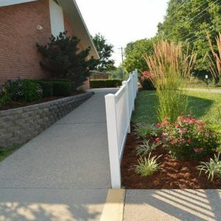 A well-maintained garden alongside a concrete pathway with a white fence next to a brick building.