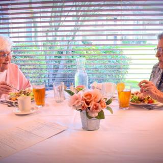 Two elderly people enjoying a meal together at a table with flowers and drinks.