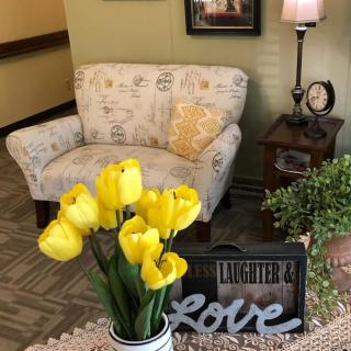 A cozy living room corner with a patterned chair, yellow tulips in a vase, and decorative elements like a lamp, clock, and inspirational sign.