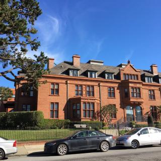 A historic brick mansion with multiple chimneys and dormer windows, set against a clear blue sky, with parked cars in front.