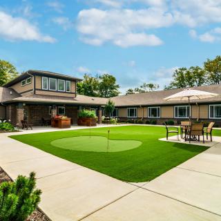 A spacious backyard featuring a putting green, surrounded by a modern house with patio seating and landscaped greenery.