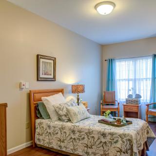 A cozy and well-lit bedroom featuring a single bed with floral bedding, a lamp, and natural light streaming through the window.