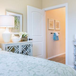 A cozy bedroom featuring elegant decor with a lamp on a mirrored dresser and an open door leading to a hallway with towels and artwork displayed.