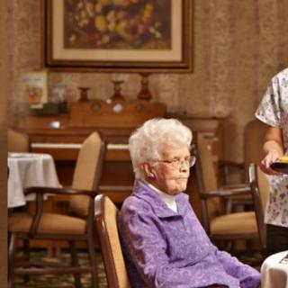 An elderly woman sitting in a care home, attended by a staff member.
