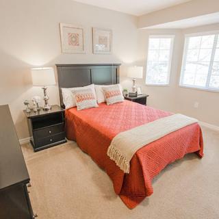 A neatly arranged bedroom with a double bed featuring an orange bedspread, two matching nightstands with lamps, and bright windows allowing natural light.