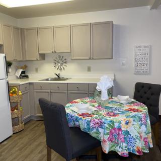 A cozy kitchen and dining area featuring a table with a colorful floral tablecloth, surrounded by chairs, with cabinets and kitchen appliances in the background.