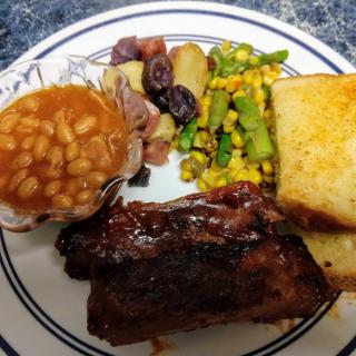 A dinner plate featuring ribs, beans, roasted potatoes, corn and asparagus mix, and a slice of bread.