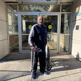 An elderly person stands outside a building, using a walker for support.