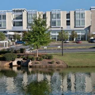 A modern building with large windows is reflected in a nearby lake, surrounded by trees and greenery on a sunny day.