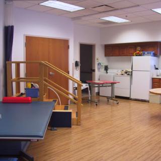 A well-equipped rehabilitation therapy room featuring exercise tables, a staircase for mobility training, and a kitchenette.