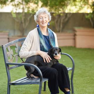A smiling woman sitting on a bench with her dog in a serene outdoor setting.
