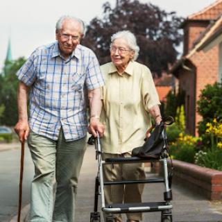 An elderly couple enjoys a walk together on a sunny day, with the woman using a walker for support.