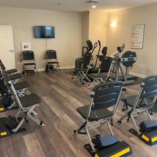 A small fitness room equipped with exercise machines, chairs for workout sessions, and a TV screen on the wall.