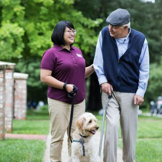 An elderly man and a caregiver walking a dog in the park.