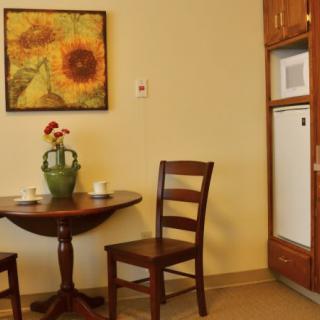 A cozy kitchen corner with a small round table, two wooden chairs, a vase with flowers, and sunflower artwork on the wall.