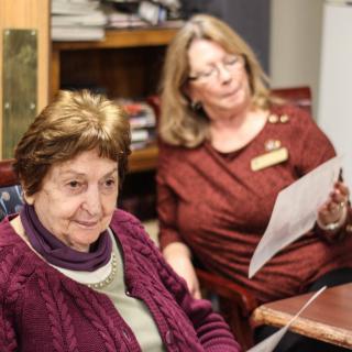 Two women sit indoors, one reading a document with a bookshelf in the background.