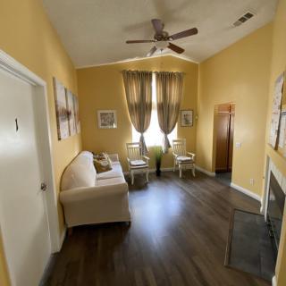A cozy living room with a sofa, curtains, and a ceiling fan, featuring warm yellow walls and wooden flooring.
