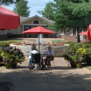 Two people sitting under red umbrellas on a patio near a garden and fountain.