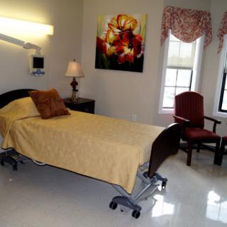 A well-lit hospital room featuring a single bed with yellow bedding, a comfortable chair, and decorative artwork on the wall.