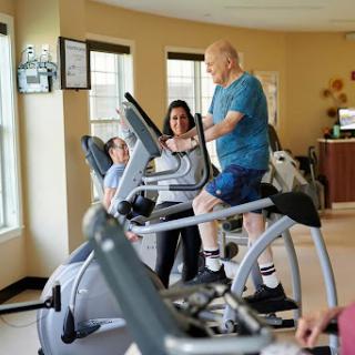 An elderly man engages in a workout on an elliptical machine with the guidance of a personal trainer at a fitness center.