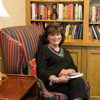 A woman sits comfortably in an armchair with a book, smiling in front of a bookshelf.