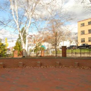 A small park area with a brick-paved ground, surrounded by trees and fenced off from nearby buildings under a partly cloudy sky.