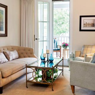 A cozy living room featuring a comfortable sofa and armchair, with a glass-top coffee table adorned with decorative items, all lit by natural light from the open balcony door.