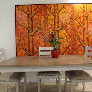 A dining room setup with a wooden table and chairs, adorned with a vibrant orange painting in the background and a potted plant on the table.