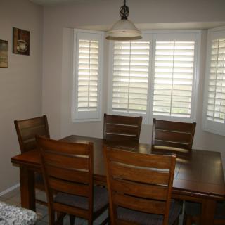 A cozy dining area with a wooden table and four chairs, complemented by large bay windows with shutters.