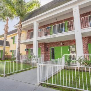 A two-story hotel building with green doors and balconies, surrounded by palm trees and a white fence.