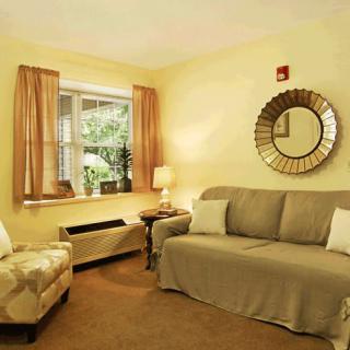 A cozy living room with a sofa, armchair, and mirror, featuring warm lighting from a lamp.