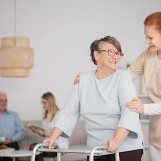 A caregiver assisting an elderly woman using a walker, both smiling in a warm and friendly environment.