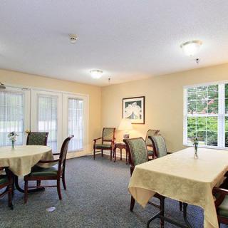 A cozy dining room with tables and chairs set for a meal, featuring soft lighting and large windows.