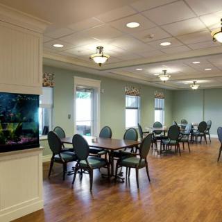 A well-lit dining area with multiple tables and chairs, featuring a television on the wall and wooden flooring.