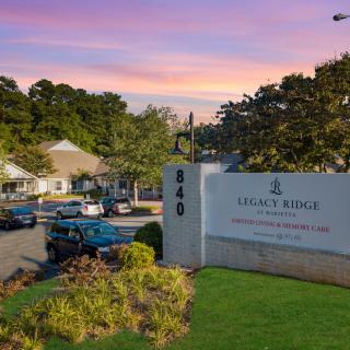 A view of Legacy Ridge, a retirement community, with its sign in the foreground and building structures surrounded by trees at dusk.
