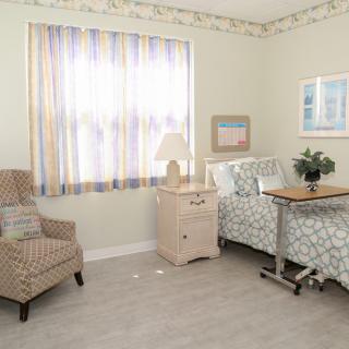 A cozy bedroom with a well-made bed, a comfortable chair, and a nightstand with a lamp by the window.