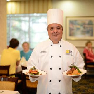 A chef in a professional kitchen holding two plates of food with a smile.