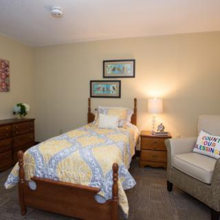 A cozy bedroom featuring a single bed with colorful bedding, a nightstand with a lamp, and an armchair beside it.