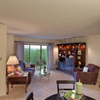 A cozy living room featuring comfortable seating, a coffee table, and a well-organized bookshelf, illuminated by natural light through sliding glass doors.
