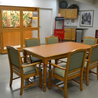 A dining area with a wooden table, surrounded by chairs, and a kitchenette in the background.
