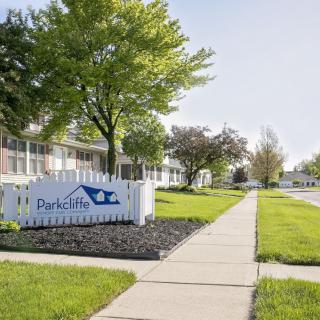 A peaceful residential area with apartments, a well-maintained lawn, and a sidewalk lined with trees.