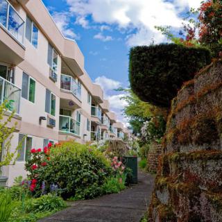A scenic view of apartment buildings with a pathway lined by lush greenery and flowers, under a partly cloudy blue sky.
