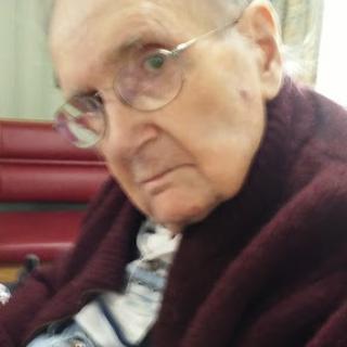 An elderly man wearing glasses and a sweater, sitting indoors.