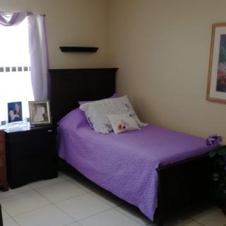 A cozy bedroom with a single bed and purple bedding, complemented by a nightstand with picture frames and a window letting in natural light.