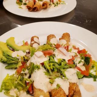 A plate of taquitos garnished with crema, fresh vegetables, and slices of avocado.
